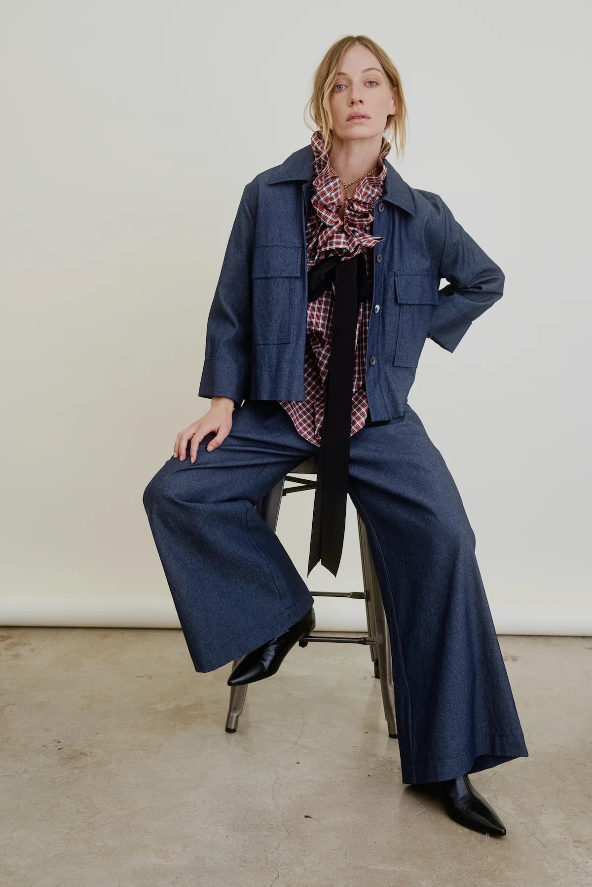 A woman confidently poses on a stool against a plain background, wearing the Aquarius Cocktail Connor Wide Leg Pant with an elastic waist, paired with a matching denim jacket, plaid ruffled blouse with black tie, and black boots.