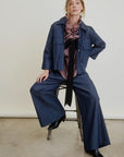 A woman confidently poses on a stool against a plain background, wearing the Aquarius Cocktail Connor Wide Leg Pant with an elastic waist, paired with a matching denim jacket, plaid ruffled blouse with black tie, and black boots.