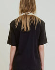 A person with long, light brown hair stands with their back to the camera against a plain light background, wearing the oversized black Campbell OS Crew Tee by Aquarius Cocktail and a layered pearls and gold chain necklace.