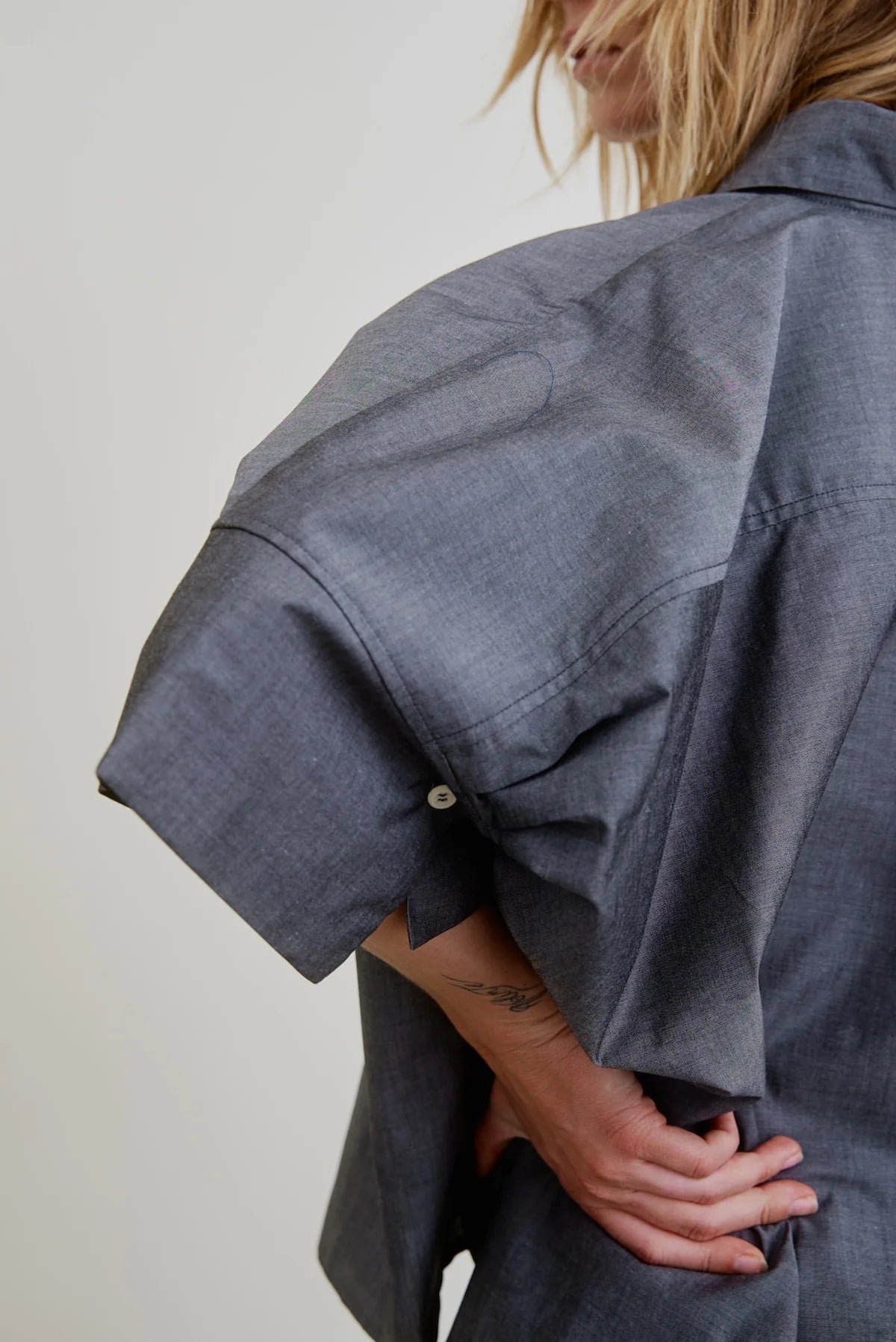 A person is seen from the side wearing the Aquarius Cocktail Vesper OS Shirt— a loose-fitting, gray chambray cotton top with short cuffed sleeves— their hand on their hip, part of their face and blonde hair visible.