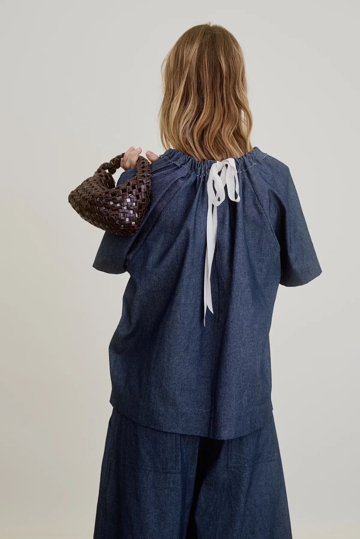 Seen from behind, a person with wavy, shoulder-length hair wears the Aquarius Cocktail Spritz Short Sleeve Gathered top in Japanese cotton denim, featuring a feminine neckline and white back ribbon tie, paired with matching blue bottoms and a dark woven bag.