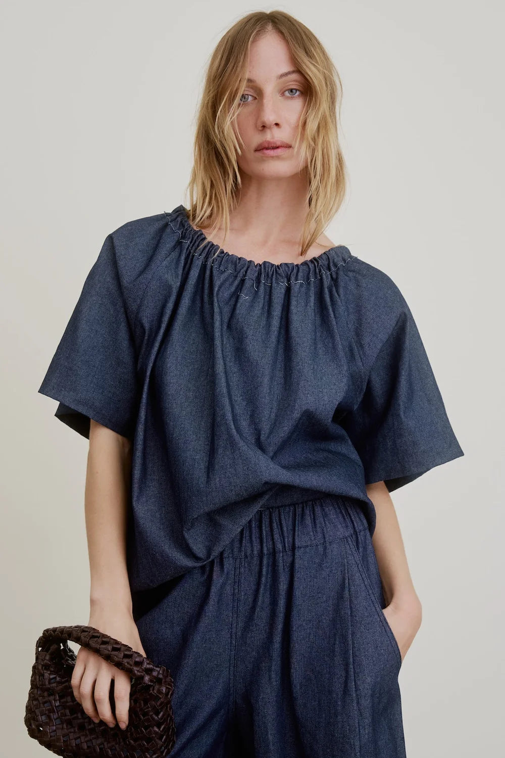 A woman with blonde, wavy hair wears the Aquarius Cocktail Spritz Short Sleeve Gathered top and matching pants in navy Japanese cotton denim, holding a small woven brown bag against a light background.