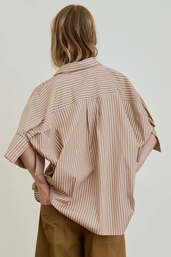 A person with shoulder-length light brown hair is seen from behind wearing the Aquarius Cocktail Vesper OS Shirt, a loose-fitting beige and white striped cotton blouse, paired with brown pants against a plain light background.