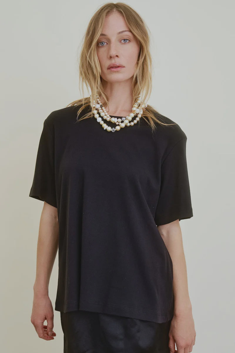 A person with long blonde hair wears the oversized black Campbell OS Crew Tee by Aquarius Cocktail, a black skirt, and layered pearl necklaces, standing against a plain light-colored background.
