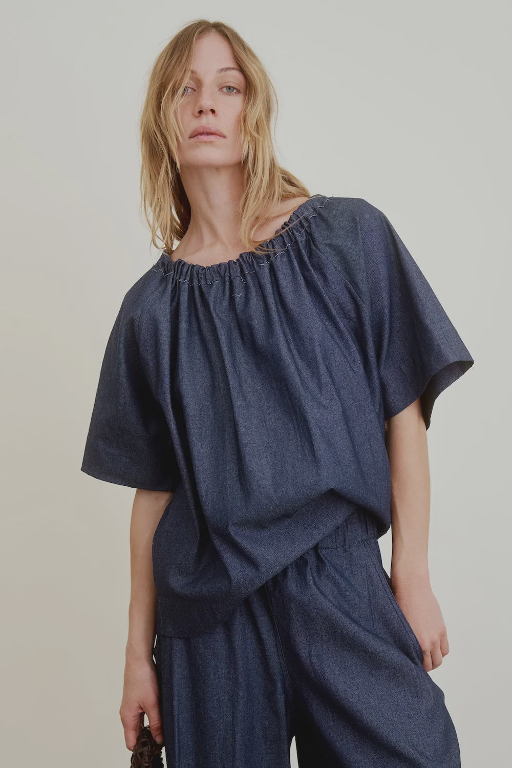 A person with long blonde hair wears the Aquarius Cocktail Spritz Short Sleeve Gathered top and matching wide-leg pants, styled loosely and off-shoulder, posing against a plain light background.