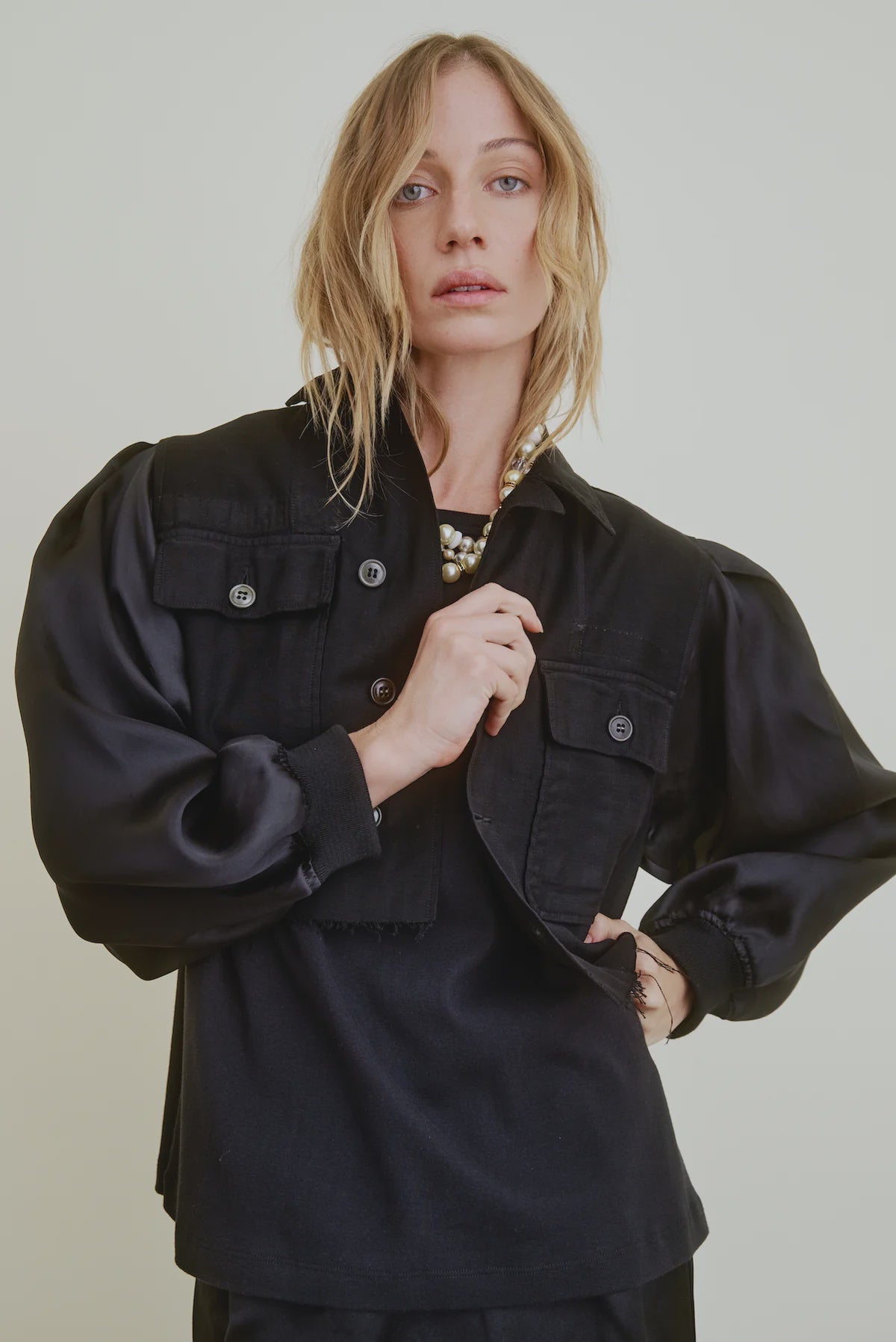 A woman with wavy blonde hair models the Ava Cropped Jacket Organza Sleeve by Aquarius Cocktail, featuring silk organza sleeves and large front pockets, styled with a chunky necklace against a plain light background.
