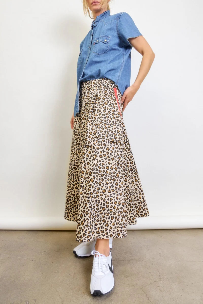 A woman stands against a plain background wearing the Aquarius Cocktail Le Cargo Skirt, styled with a short-sleeved denim shirt and white sneakers. Her face is partially out of frame.