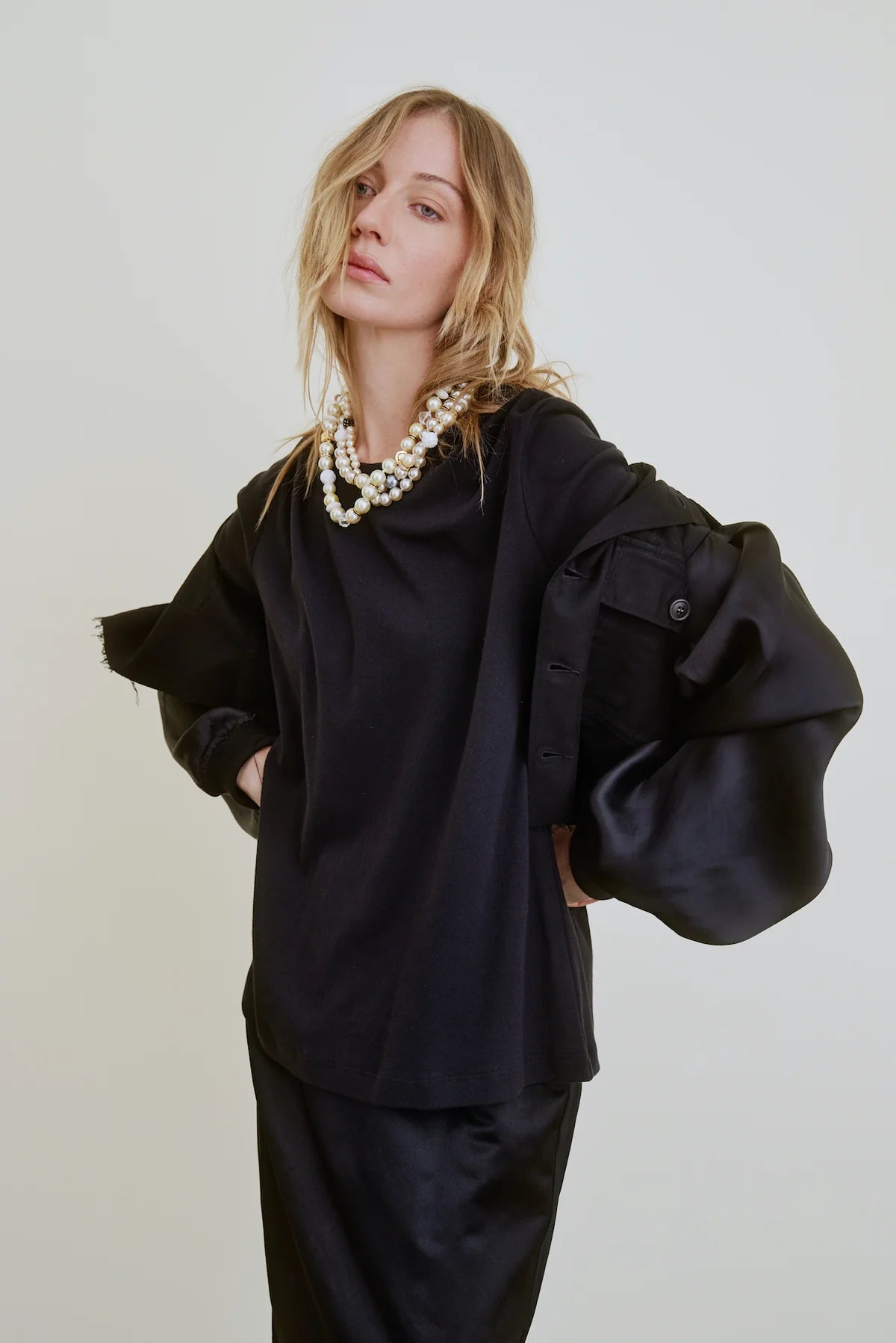 A woman with wavy blonde hair wears the Aquarius Cocktail Ava Cropped Jacket with organza sleeves and a layered pearl necklace, standing against a plain light background.