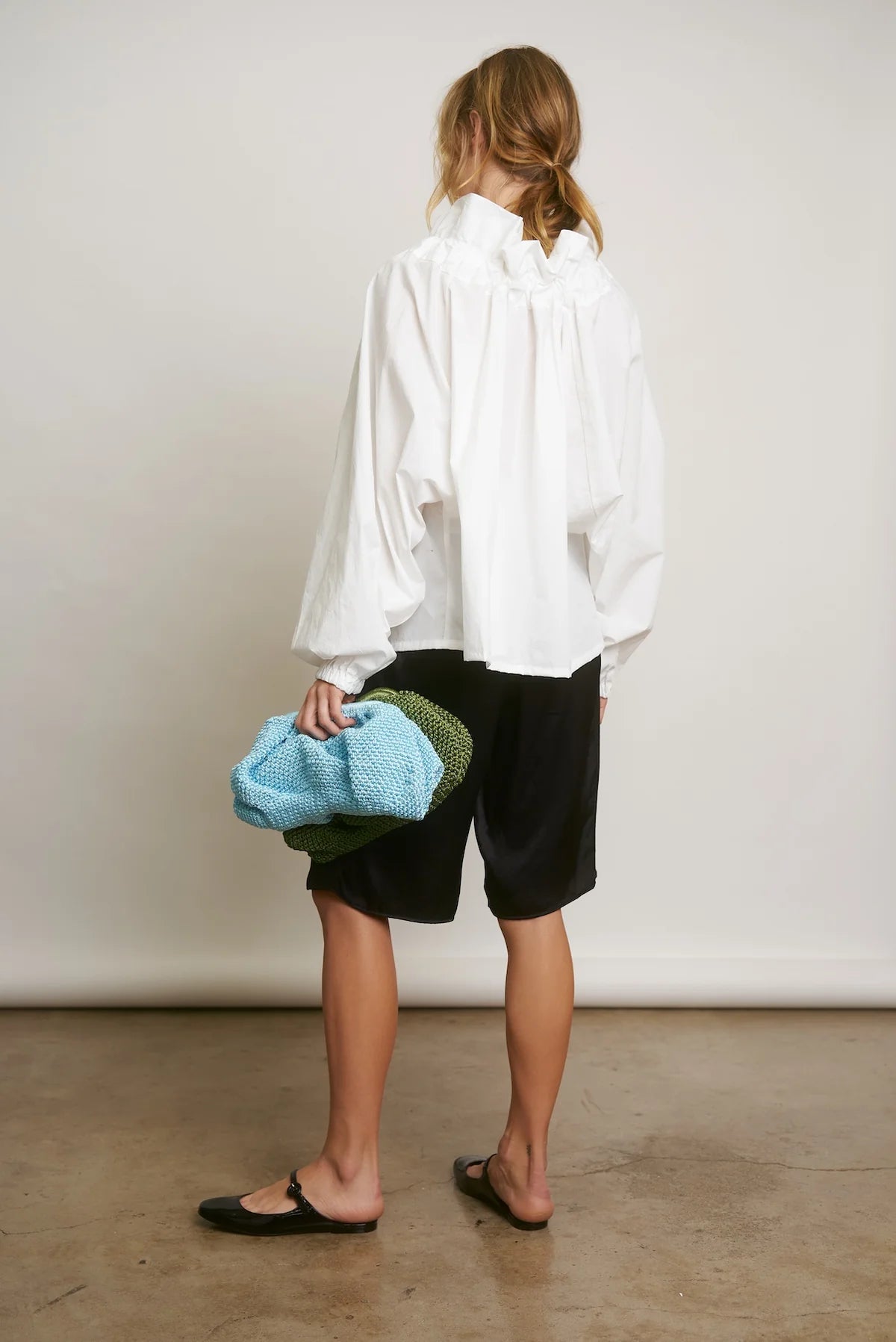 Facing away, the person wears the Aquarius Cocktail Beautiful Mess Poet Blouse with black knee-length shorts and black flats, holding a textured blue and green bag in their left hand against a plain background.