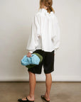Facing away, the person wears the Aquarius Cocktail Beautiful Mess Poet Blouse with black knee-length shorts and black flats, holding a textured blue and green bag in their left hand against a plain background.