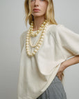 A woman with long blonde hair wears the Aquarius Cocktail Ruth French Terry SS Sweatshirt in cream, layered pearl necklaces, and gray pants, posing with her hand on her hip against a light gray background.