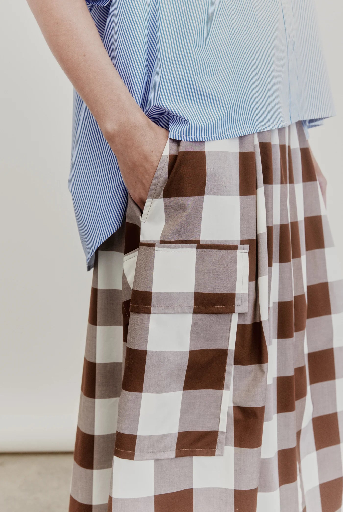 Wearing the Aquarius Cocktail Le Cargo Skirt, crafted from Japanese cotton poplin in a brown-and-white checkered pattern, a person stands with one hand tucked into one of the skirt’s oversized cargo pockets, paired with a blue striped top.