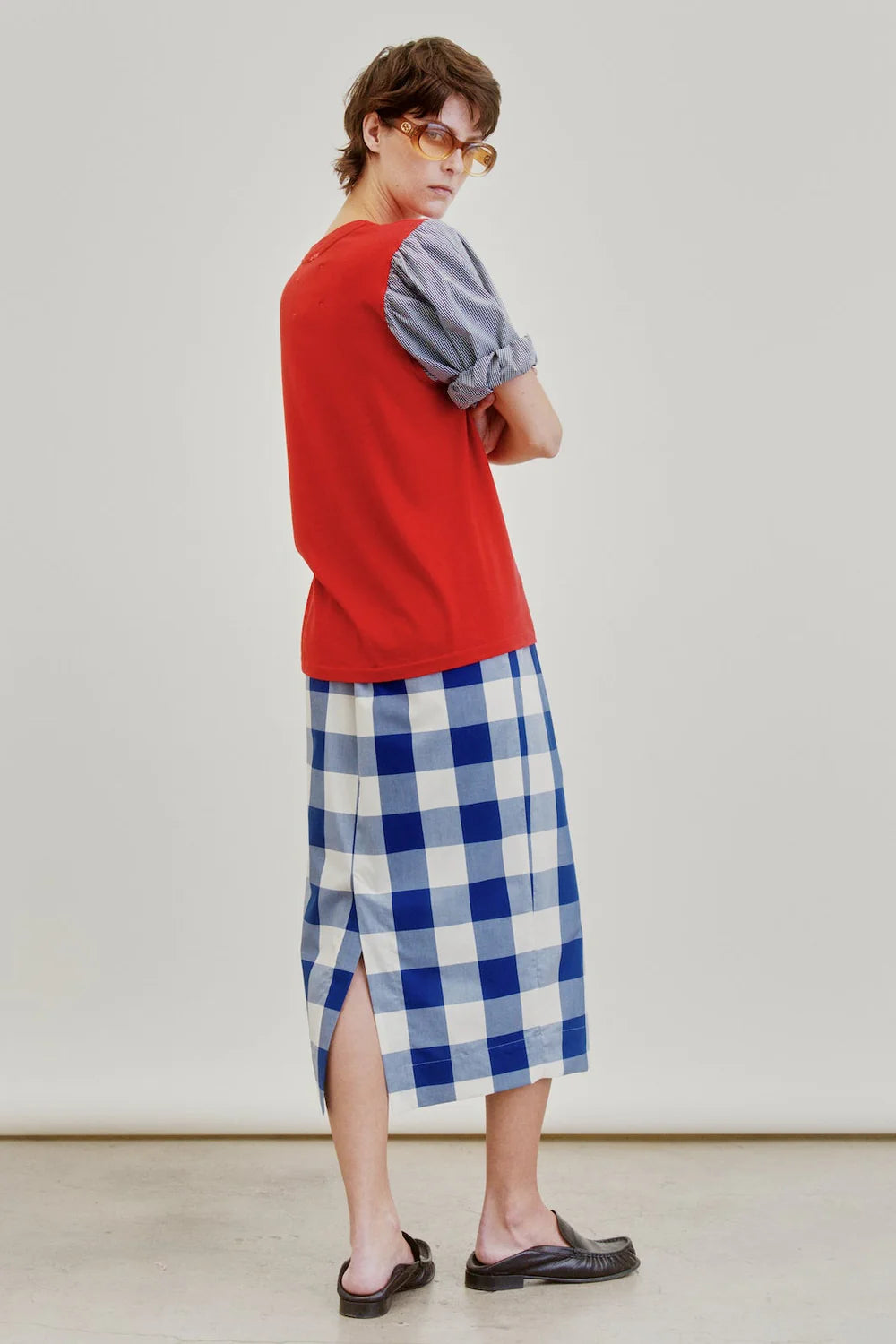 A person wearing amber sunglasses, a red sleeveless top over a gray shirt, and the Aquarius Cocktail Le Pencil Slim Skirt in blue-and-white Japanese cotton poplin stands in black sandals, arms crossed, looking over their shoulder.