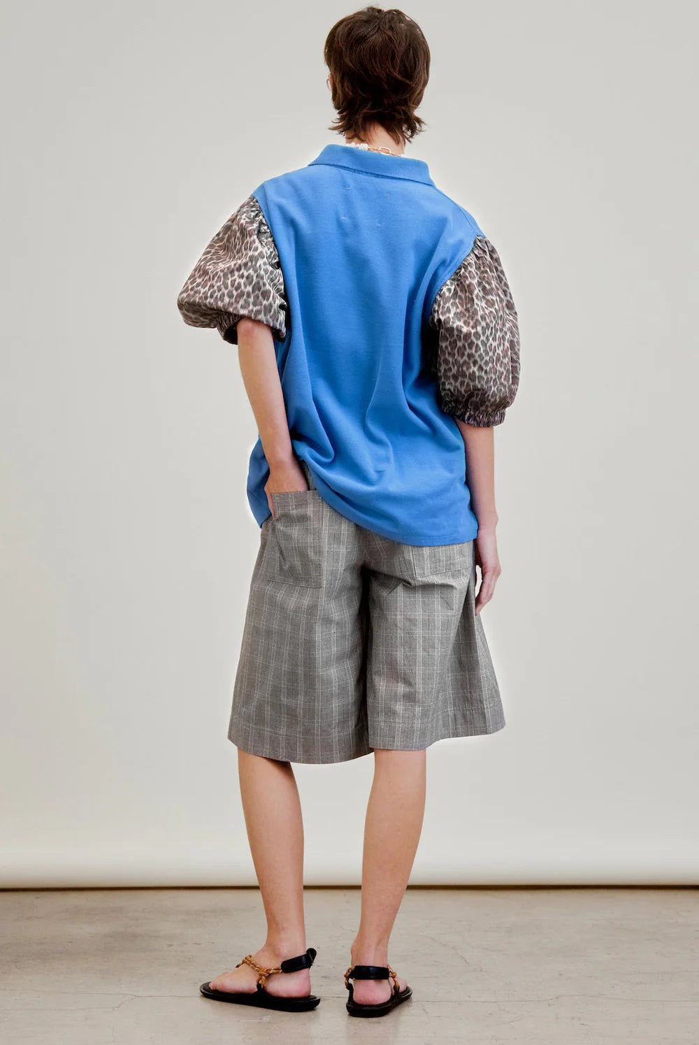 A person stands with their back to the camera in a blue top with puffed, leopard print sleeves, black sandals, and Aquarius Cocktail's Coupe Knee Length Short, one hand in their pocket against a plain background.