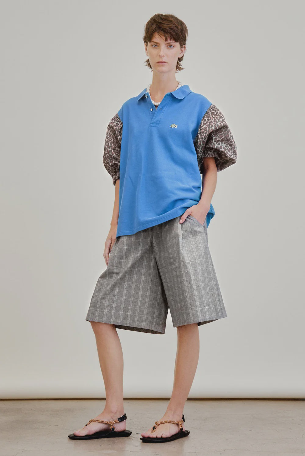 A person with short brown hair and a neutral expression stands against a plain backdrop wearing a blue polo with leopard-print puffed sleeves, black sandals with gold chain straps, and Aquarius Cocktail's Coupe Knee Length Short.