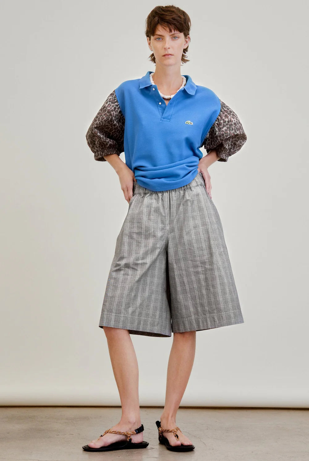 A person stands with hands in pockets, wearing a blue polo shirt, Aquarius Cocktail Coupe Knee Length Short, and black sandals. The background is plain and neutral.