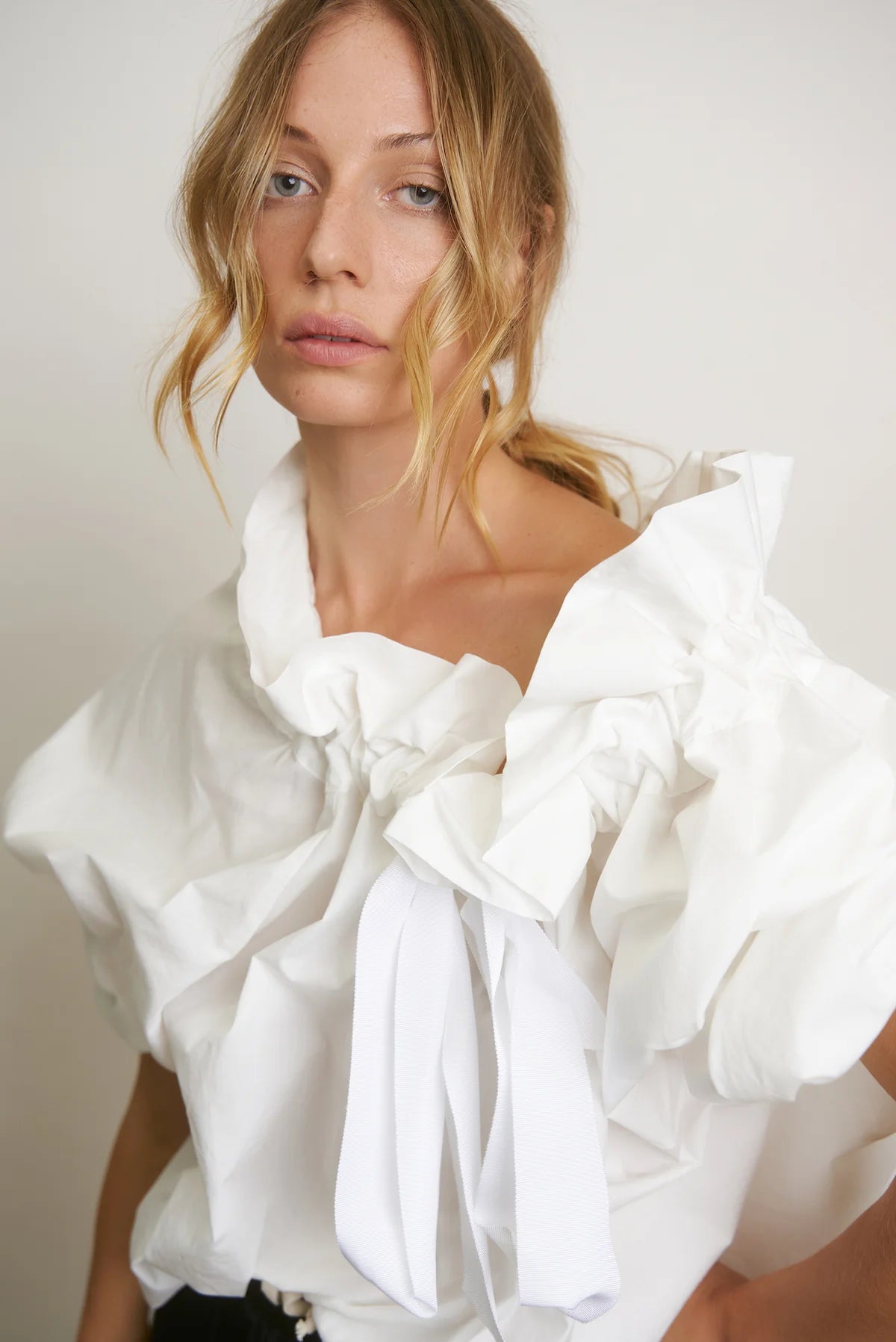 A woman with wavy blonde hair wears the Beautiful Mess Poet Blouse by Aquarius Cocktail, featuring sculptural ruffles and white ribbon accents, standing against a plain light background.