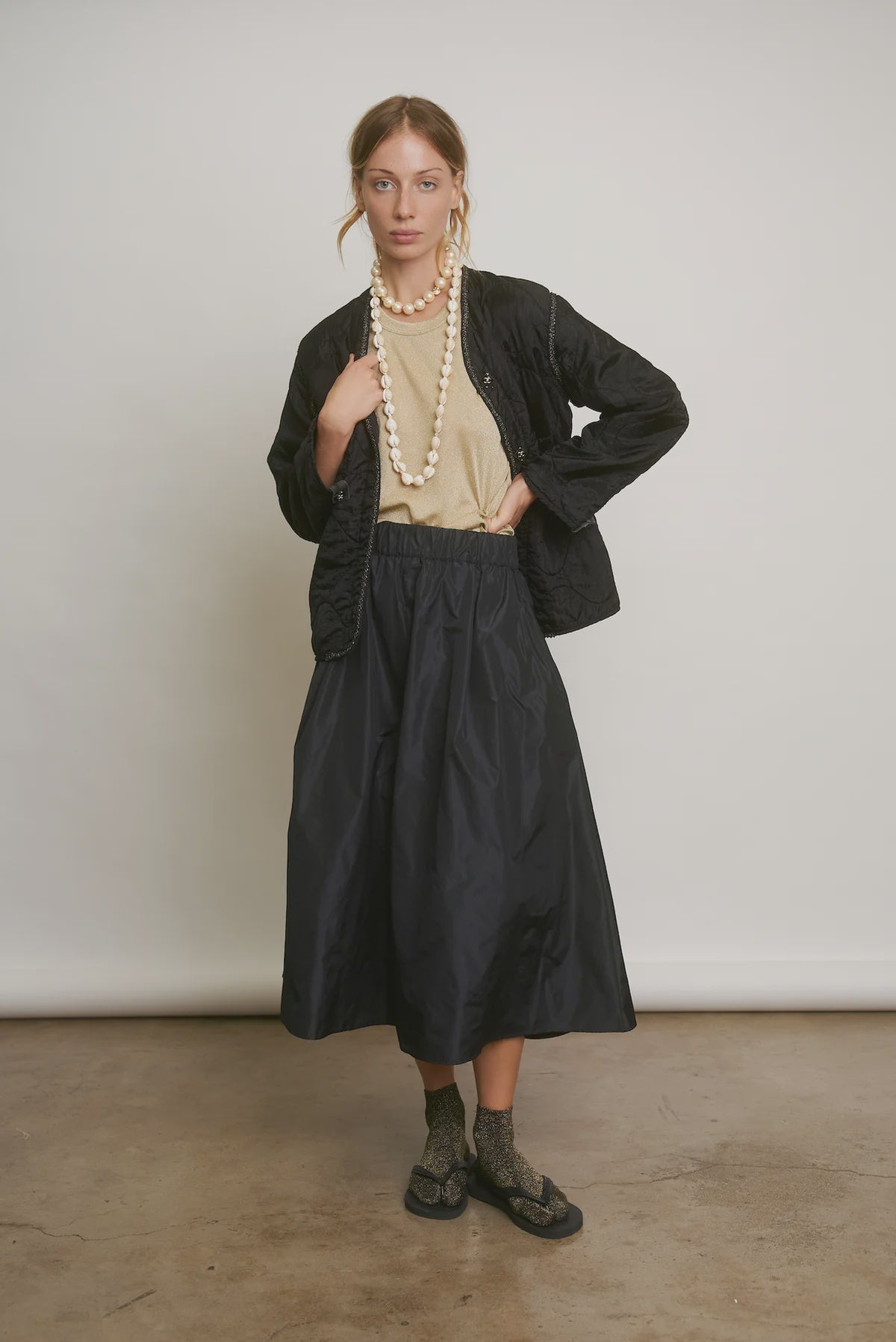 A person stands against a plain background wearing a black quilted jacket, beige top, and the LE SKIRT Silk Taffeta from Aquarius Cocktail. They accessorize with a long pearl necklace, gray socks, and black sandals. Their hair is pulled back as they pose with one hand on their jacket.