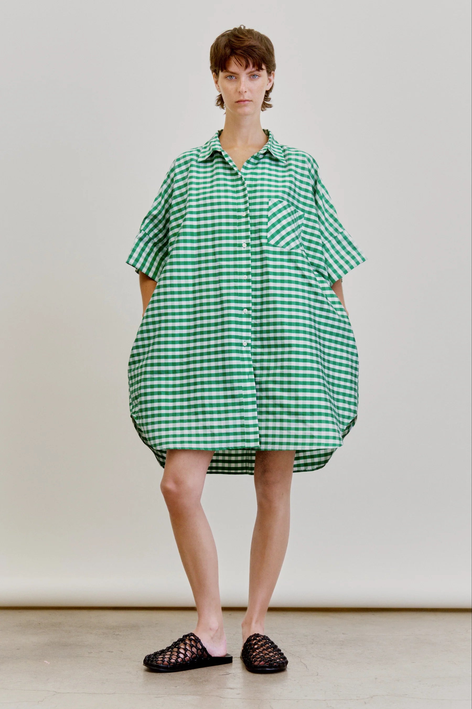 A person stands before a plain background wearing the oversized Ging Bellini Dress by Aquarius Cocktail, featuring green and white gingham with short sleeves, paired with black woven slip-on shoes.