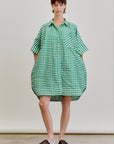 A person stands before a plain background wearing the oversized Ging Bellini Dress by Aquarius Cocktail, featuring green and white gingham with short sleeves, paired with black woven slip-on shoes.