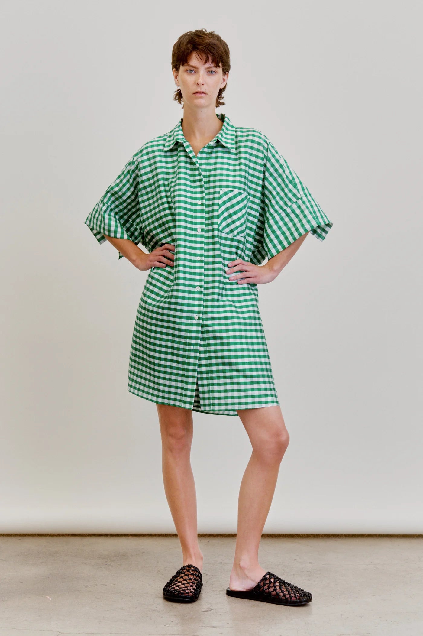 A person stands with hands on hips, wearing the oversized green and white Aquarius Cocktail Ging Bellini Dress and black slip-on woven shoes, set against a plain light background.