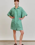 A person stands with hands on hips, wearing the oversized green and white Aquarius Cocktail Ging Bellini Dress and black slip-on woven shoes, set against a plain light background.
