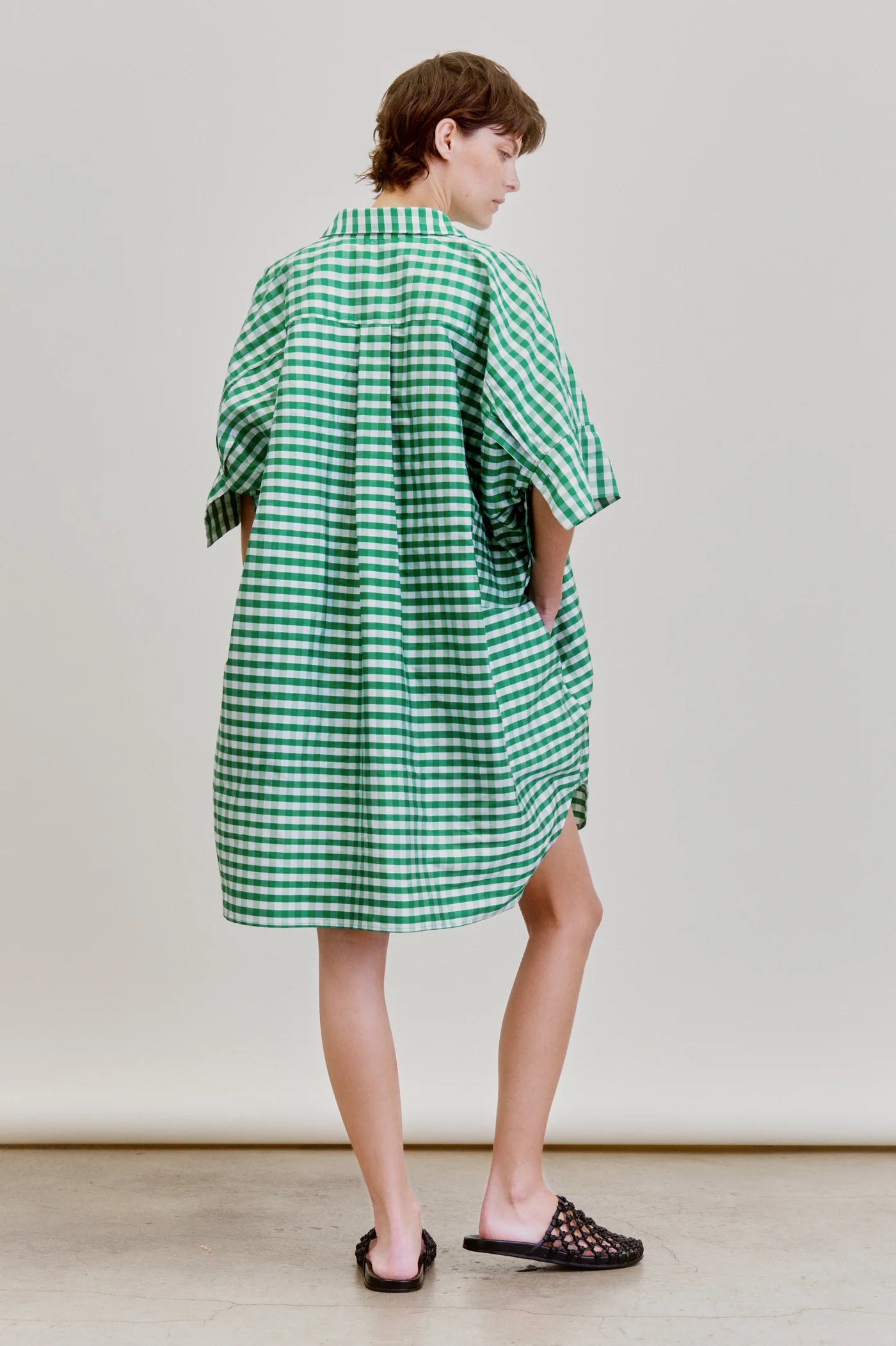 A person faces away from the camera, wearing the Aquarius Cocktail Ging Bellini Dress—an oversized green and white gingham shirt dress—and black woven slide sandals, set against a plain light background.
