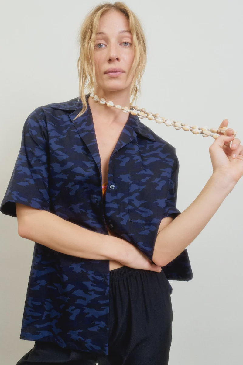 A blonde woman wears the Aquarius Cocktail CAMO LE Camp Shirt—a blue camouflage-patterned, boxy-fit style—paired with black pants, holding a shell necklace against a plain light background.