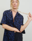 A blonde woman wears the Aquarius Cocktail CAMO LE Camp Shirt—a blue camouflage-patterned, boxy-fit style—paired with black pants, holding a shell necklace against a plain light background.