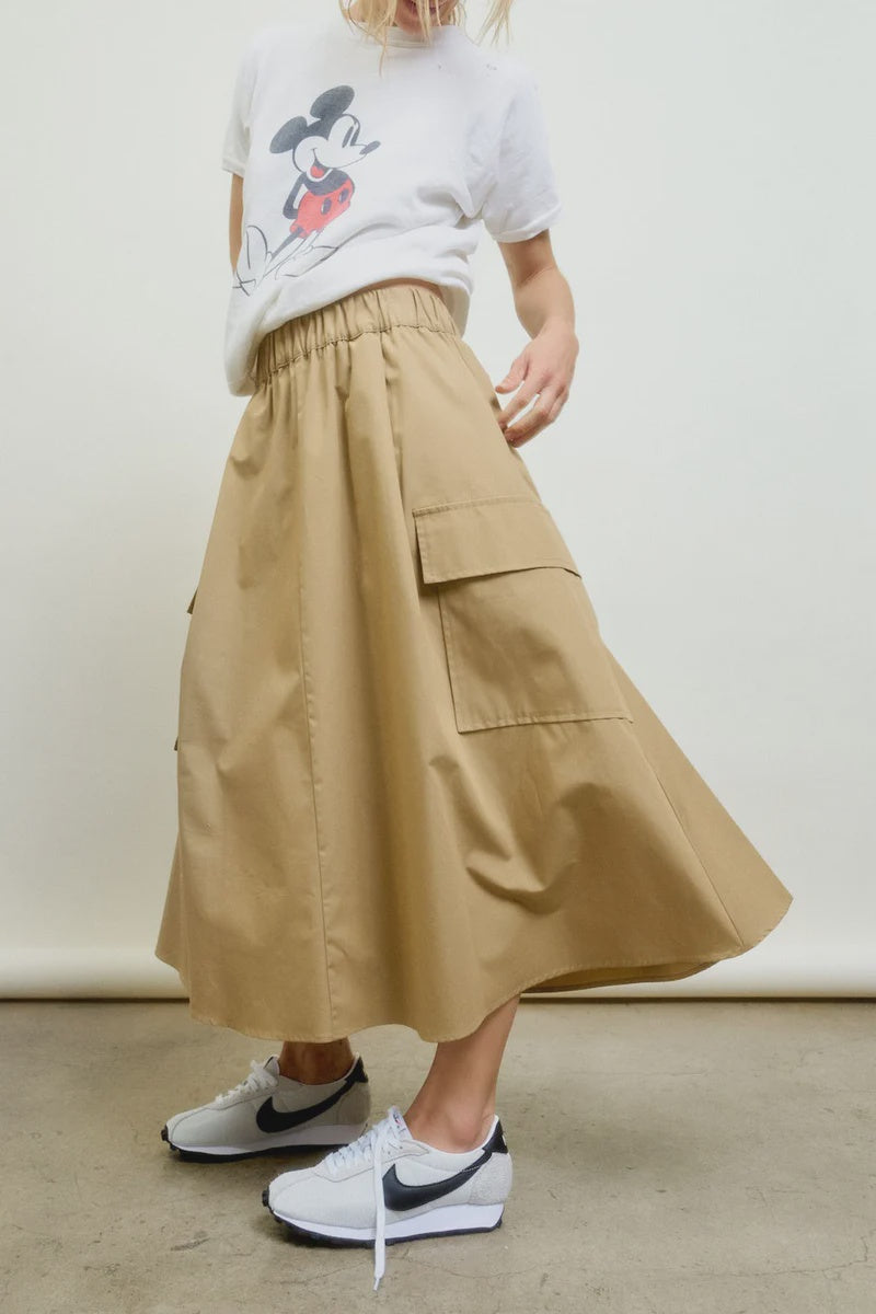 A person wears a white Mickey Mouse graphic tee with the Le Cargo Skirt by Aquarius Cocktail, featuring oversized pockets and Japanese cotton, paired with white Nike sneakers against a plain light background.