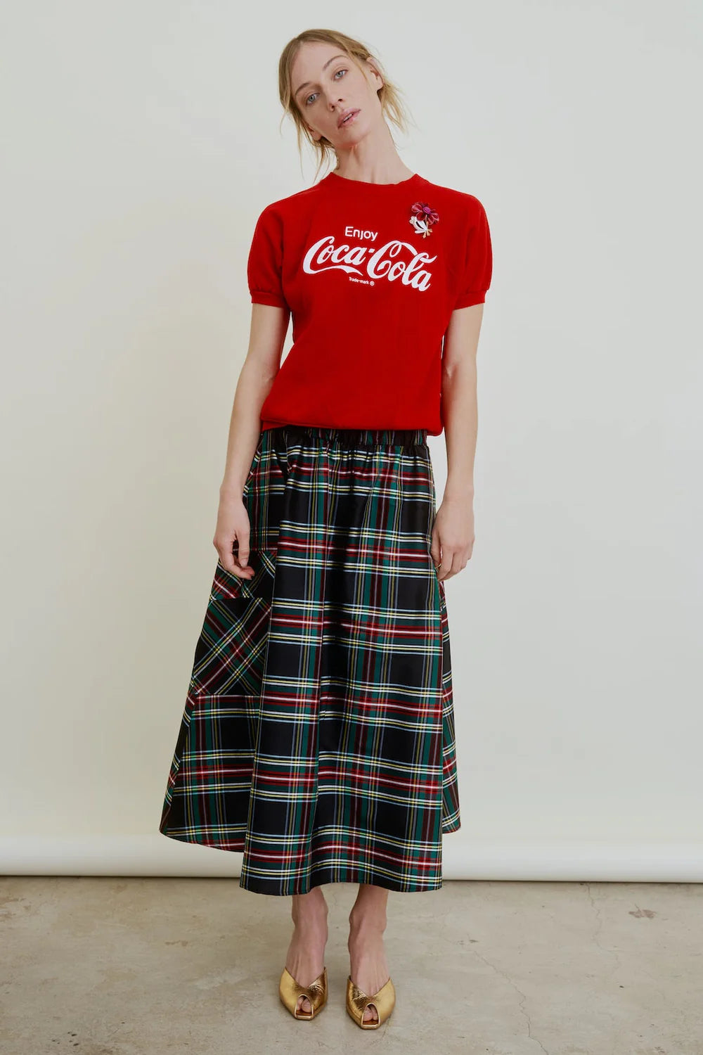 A woman stands against a plain background wearing a red Coca-Cola T-shirt, shiny gold flats, and the Le Cargo Skirt by Aquarius Cocktail—a long Japanese cotton poplin plaid skirt with red and green lines. She gazes at the camera, head slightly tilted.