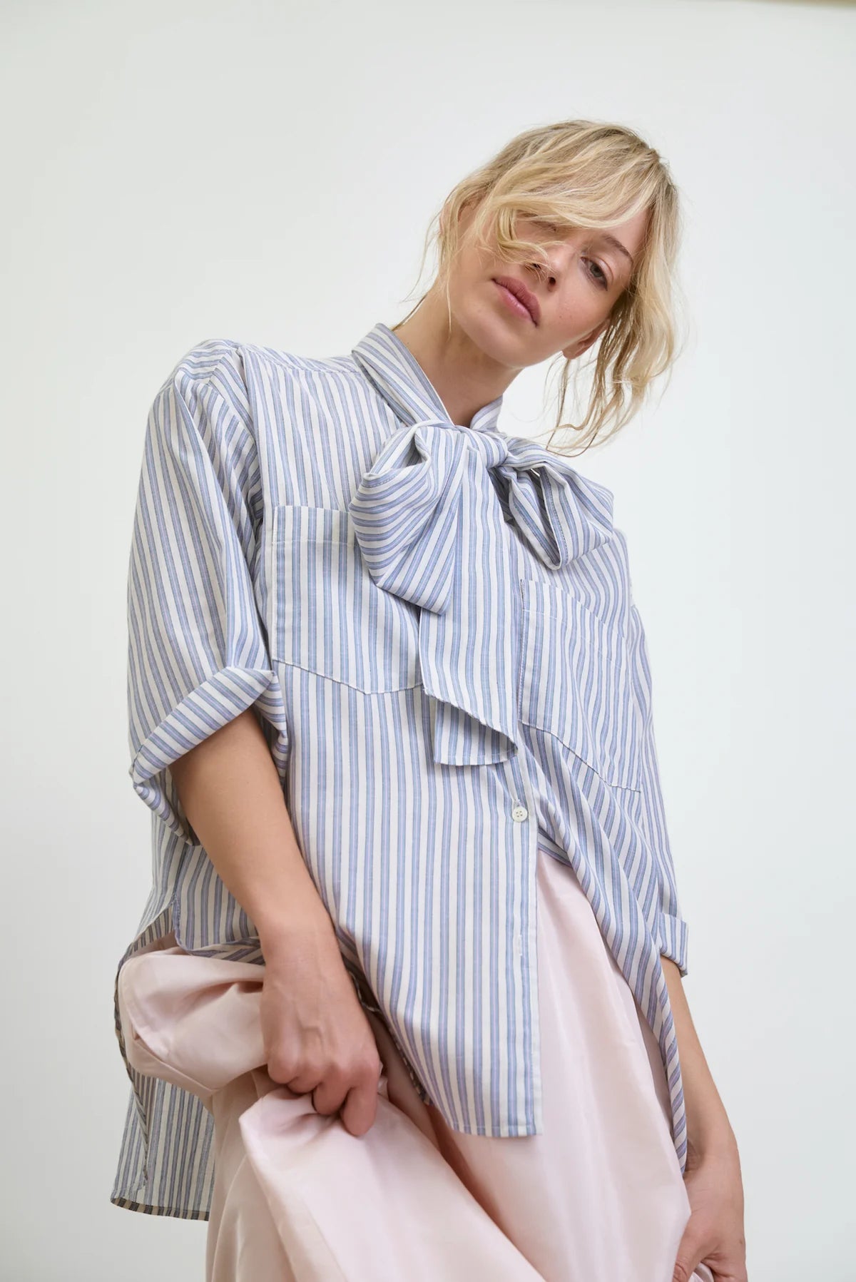 The individual wears an Aquarius Cocktail Dear Polly Shirting Bow Blouse in light blue and white with a large collar bow, paired with a pale pink skirt. Their blonde hair is styled in a loose updo, elegantly framing their face against the plain backdrop.