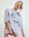 The individual wears an Aquarius Cocktail Dear Polly Shirting Bow Blouse in light blue and white with a large collar bow, paired with a pale pink skirt. Their blonde hair is styled in a loose updo, elegantly framing their face against the plain backdrop.