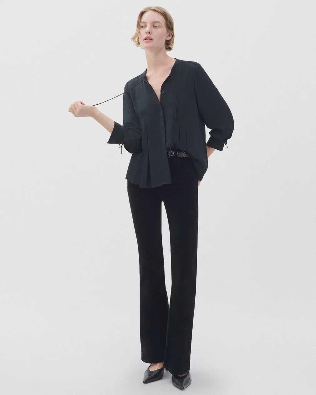 A woman stands against a plain light background wearing the Rag & Bone arden pleated blouse, styled loosely with feminine ties. She pairs it with black trousers and heels, holding one end of her blouse’s tie in her right hand.