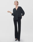 A woman stands against a plain light background wearing the Rag & Bone arden pleated blouse, styled loosely with feminine ties. She pairs it with black trousers and heels, holding one end of her blouse’s tie in her right hand.