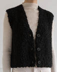 A person wears a cream ribbed turtleneck sweater with the LaTierra Aries Boucle Vest, a black, textured alpaca blend vest featuring a v neckline and three large cow horn buttons.