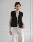 A person stands against a light gray wall wearing a cream ribbed turtleneck, high-waisted white pants, and the Aries Boucle Vest by LaTierra—a textured black alpaca blend vest with cow horn buttons.