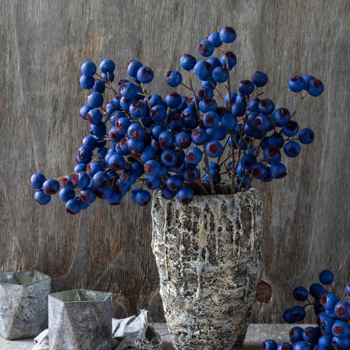 Abigail Ahern’s Hawthorn Berry Midnight features vivid blue artificial berries arranged in a rustic vase, set against a textured wooden background. Two metallic cups complement the earthy, natural decor.