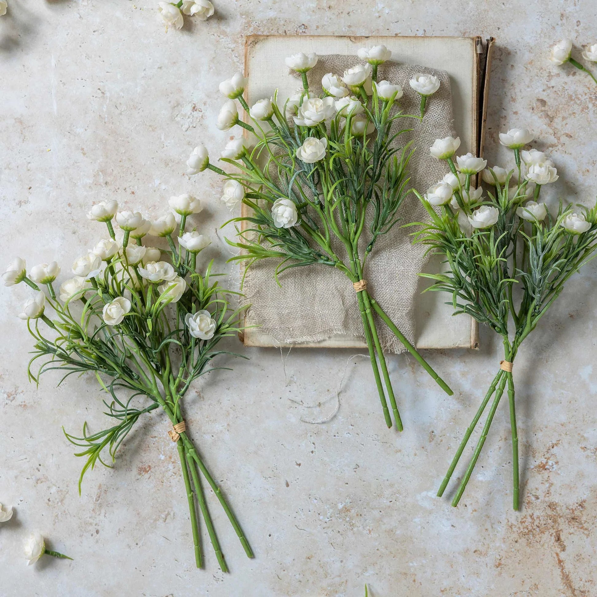 Three Hellebore Bunch, White artificial flower stems by Abigail Ahern, with green stems, are arranged on a light textured surface—one resting on a closed vintage book while others lie nearby amid scattered petals.
