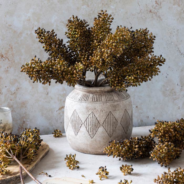 The Sorghum Stem Umber by Abigail Ahern is displayed in a rustic ceramic vase with geometric patterns, complemented by scattered seed pods and a faux floral stem on a light stone surface against a textured neutral background.
