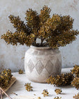 The Sorghum Stem Umber by Abigail Ahern is displayed in a rustic ceramic vase with geometric patterns, complemented by scattered seed pods and a faux floral stem on a light stone surface against a textured neutral background.
