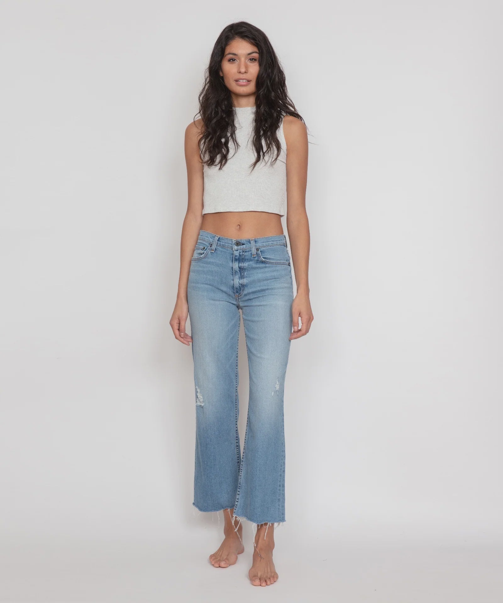 A woman with long, wavy dark hair stands against a plain white background, wearing a sleeveless, light gray cropped top paired with ASKK's Crop Wide Leg high-waisted, light blue stretch denim jeans. The jeans feature a slightly flared, frayed hem. She is barefoot and looking directly at the camera.