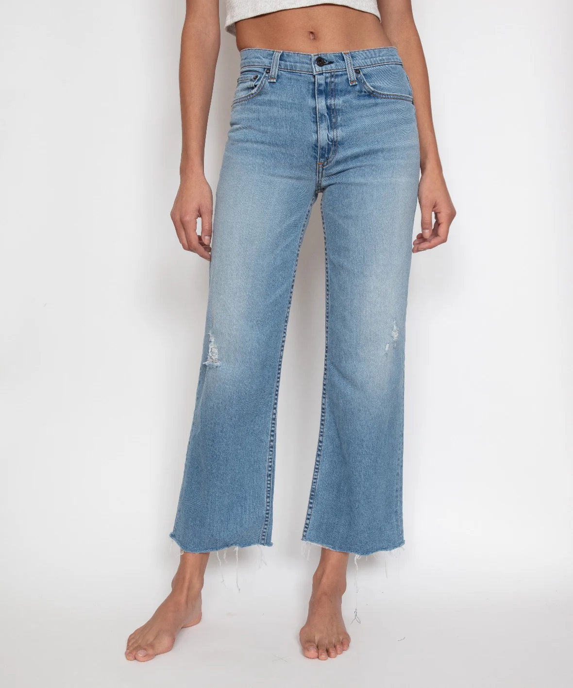 A person stands barefoot against a plain white background, dressed in ASKK's Crop Wide Leg jeans. These light blue, high-rise, wide-leg jeans are crafted from Japanese stretch denim and feature a raw, frayed hemline. The jeans display slight distressed detailing on the upper thighs and knees. The person's torso is partially visible, adorned in a cropped top.