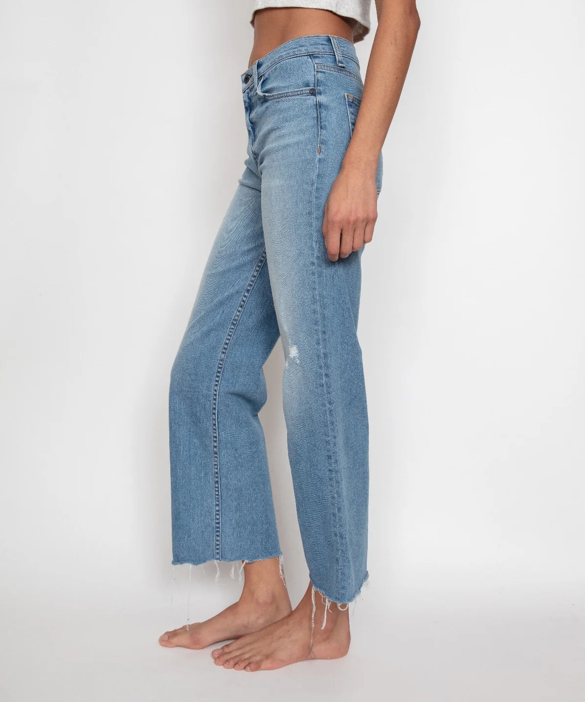 A person is modeling the Crop Wide Leg jeans by ASKK. These high-rise, light blue denim jeans with a raw hem are crafted from Japanese stretch denim and feature a straight-leg cut with subtle distressing. The cropped inseam enhances their barefoot stance as they pose in side profile against a plain white background.