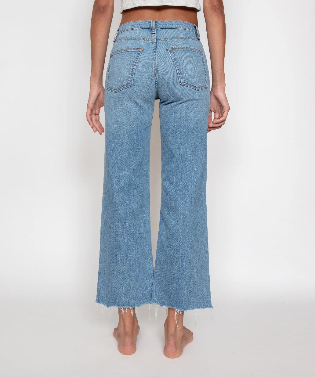 A person stands barefoot facing away from the camera, wearing the Crop Wide Leg jeans by ASKK. These high-rise, light blue high-waisted jeans feature raw, frayed hems and patch-style back pockets. Made from Japanese stretch denim, they offer a loose, wide-leg fit. The background is plain white.