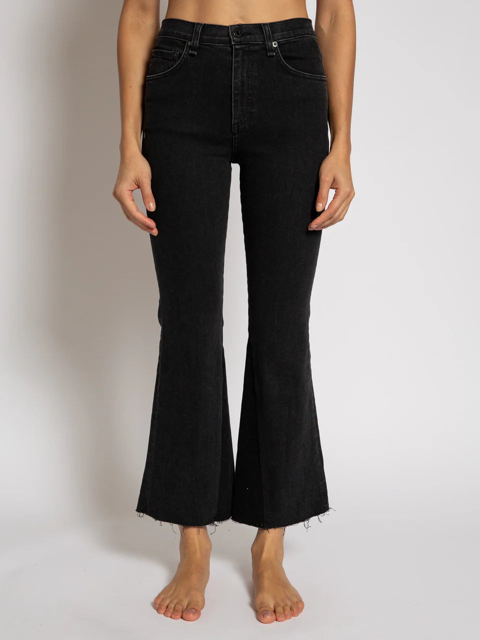 A person wearing the ASKK Cropped Trouser Boot, stylish black high-rise flared jeans, stands barefoot against a plain background. These ankle-length jeans are made from premium Japanese denim and feature distinctive frayed hems at the bottom.