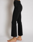 A person is wearing the Cropped Trouser Boot by ASKK, featuring black frayed-hem jeans made from Japanese denim. The high-rise design offers a snug fit and flares slightly at the ankle-length bottom. They are shown barefoot against a plain, light background.