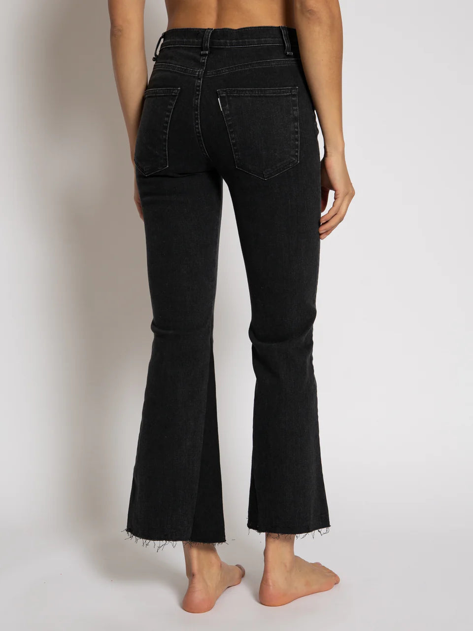 An individual stands barefoot against a plain background, wearing the Cropped Trouser Boot jeans from ASKK. These black frayed hem jeans are crafted from Japanese denim and feature a high-waisted fit with flared bottoms, shown from the back.