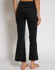 An individual stands barefoot against a plain background, wearing the Cropped Trouser Boot jeans from ASKK. These black frayed hem jeans are crafted from Japanese denim and feature a high-waisted fit with flared bottoms, shown from the back.