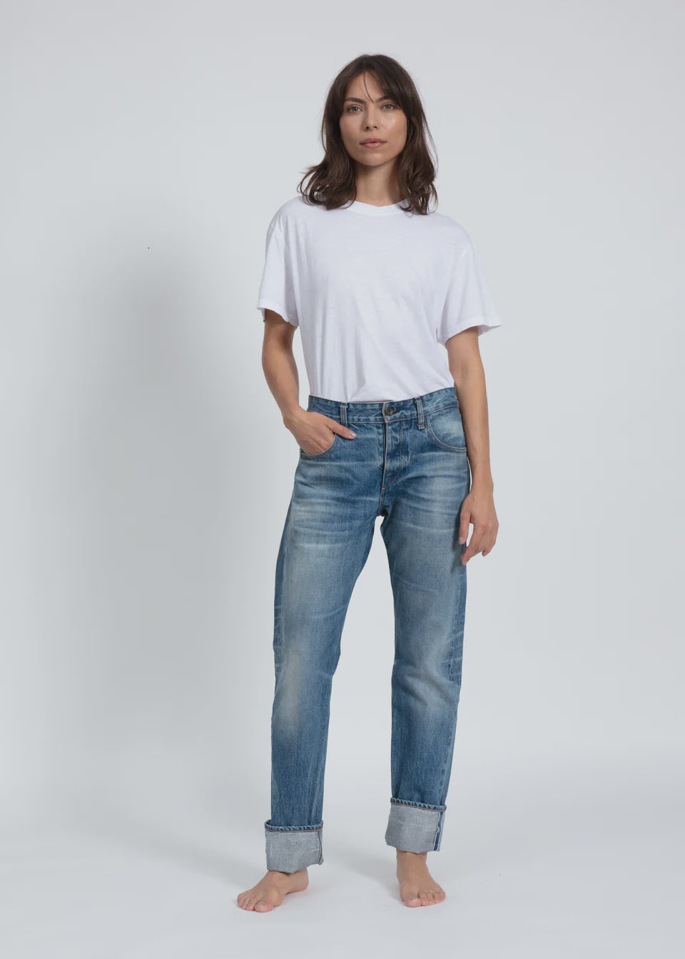 A person stands barefoot against a plain background, wearing an ASKK white t-shirt and relaxed Selvage Jean in indigo. One hand rests in the pocket while the other hangs by the side, showcasing a vintage men's style with a neutral expression.